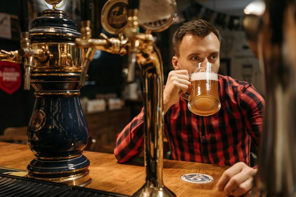 A man in a red and black checkered flannel shirt drinking a draft beer from a glass mug at a wooden bar counter, with a blue ceramic beer tap tower in the foreground.