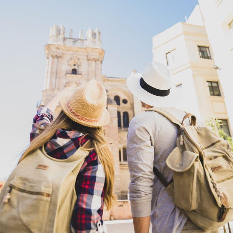 The Art of Cultural Immersion How to Travel Like a Local in Europe - Back view of a tourist couple wearing hats and backpacks, standing in front of the Málaga Cathedral and pointing up at the historic stone architecture under a clear blue sky.