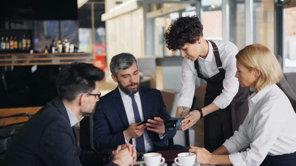 waiter holds a mobile payment terminal while a businessman in a suit pays with his smartphone at a restaurant table, accompanied by two colleagues with coffee cups in the foreground.