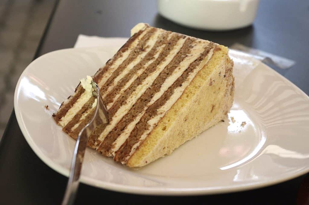 Close-up photograph of a slice of traditional Hungarian Esterházy torte on a white plate. The dessert features distinct thin layers of almond meringue dough alternating with vanilla buttercream. A silver fork is cutting into the tip of the slice, revealing the creamy texture.