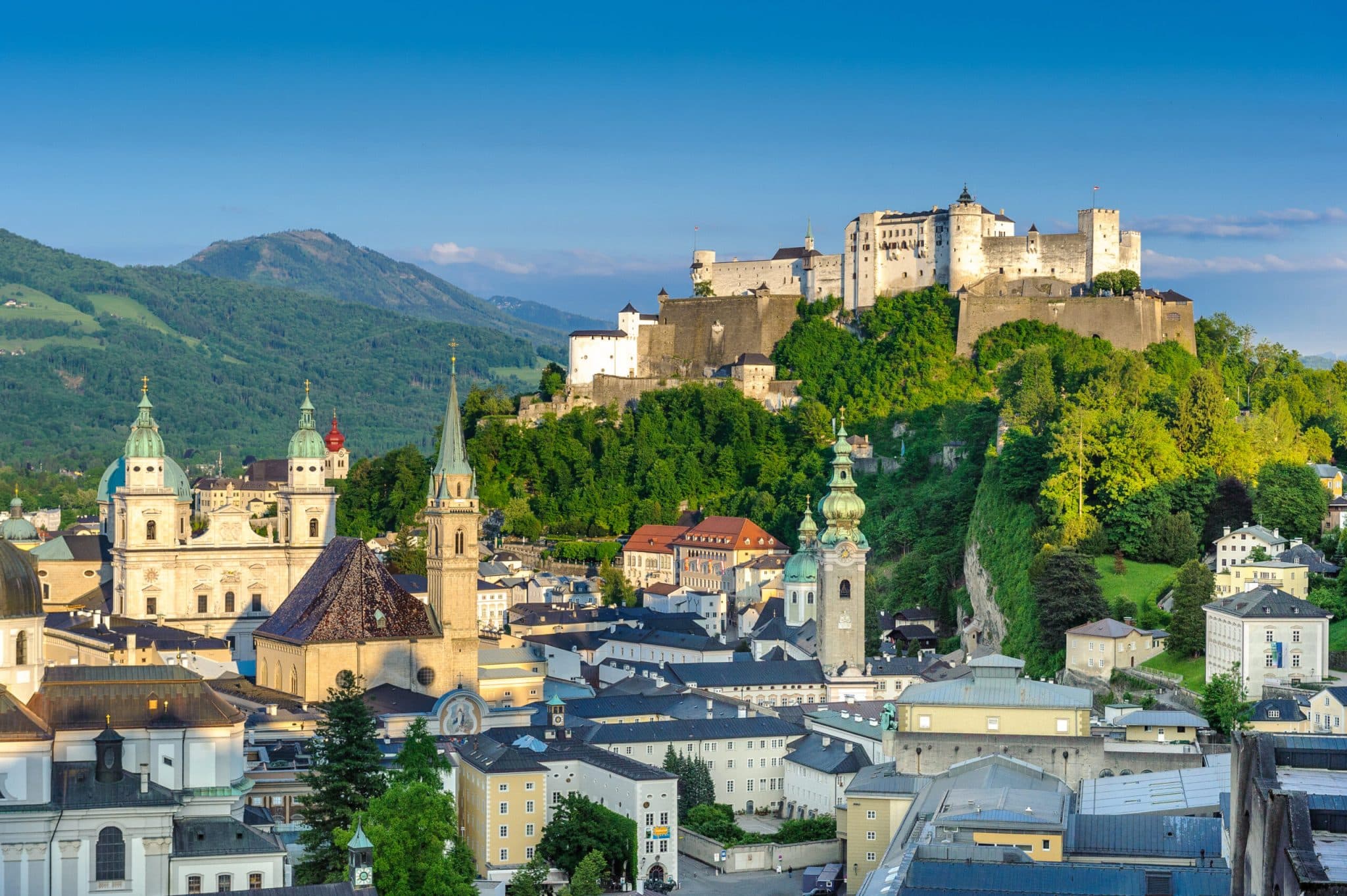 A panoramic cityscape of Salzburg, Austria, featuring the white Hohensalzburg Fortress atop a green hill overlooking the baroque Salzburg Cathedral and historic old town buildings, set against a backdrop of blue mountains.