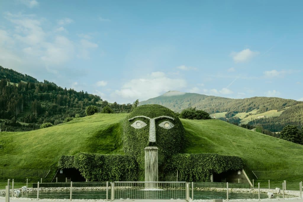 A monumental grass-covered sculpture of a giant's head embedded in a hillside at Swarovski Crystal Worlds (Kristallwelten). Water cascades from the mouth into a pool below, and the eyes are made of large sparkling crystals. The background features the Austrian Alps and a forest under a blue sky.