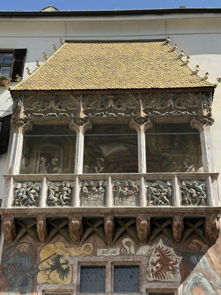 A low-angle, full view of the Goldenes Dachl (Golden Roof) in Innsbruck, Austria, featuring a balcony topped with 2,657 fire-gilded copper tiles. The facade includes intricate Gothic stone reliefs of Emperor Maximilian I and dancers, colorful frescos of standard bearers with coats of arms, and is protected by a fine wire mesh.