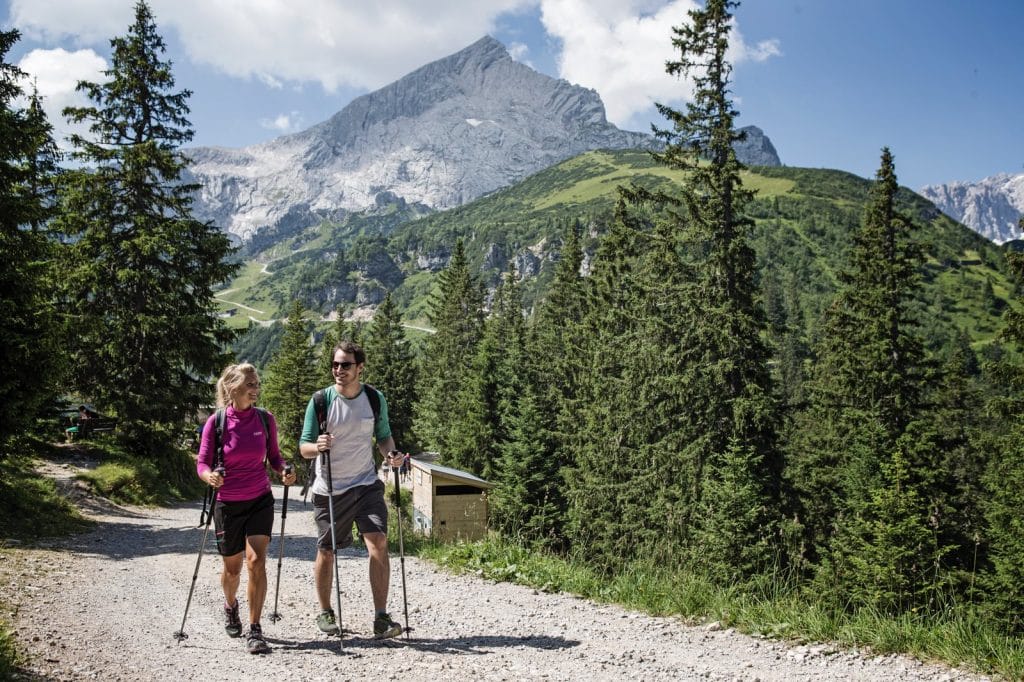 A man and a woman hiking with trekking poles on a gravel path in the summer, with the iconic Alpspitze mountain peak rising behind green pine trees in the background.