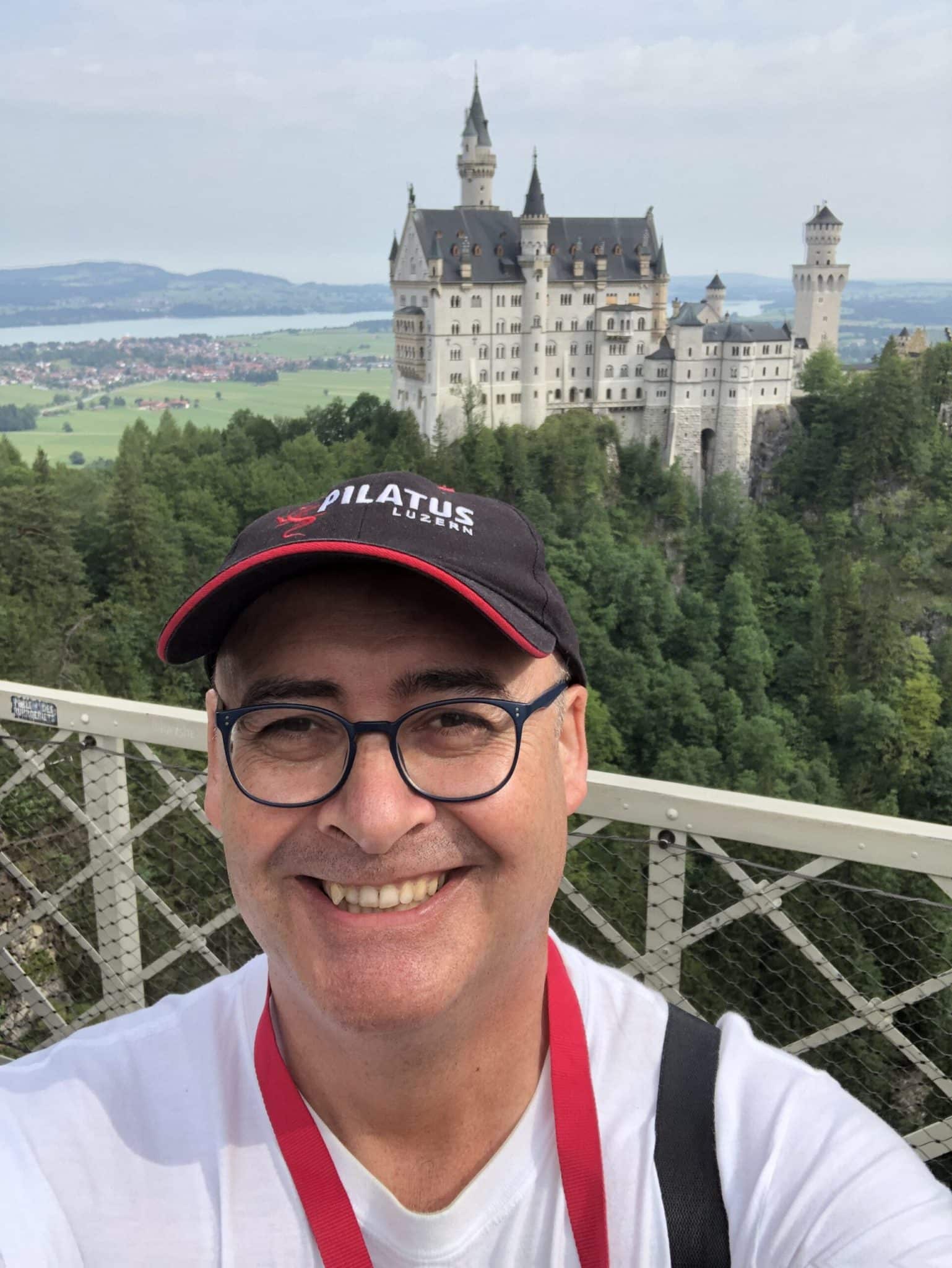 A selfie of Pieter Reynolds smiling on a bridge with Neuschwanstein Castle in the background in Germany. He is wearing glasses and a black cap with "PILATUS LUZERN" text and a red dragon logo.