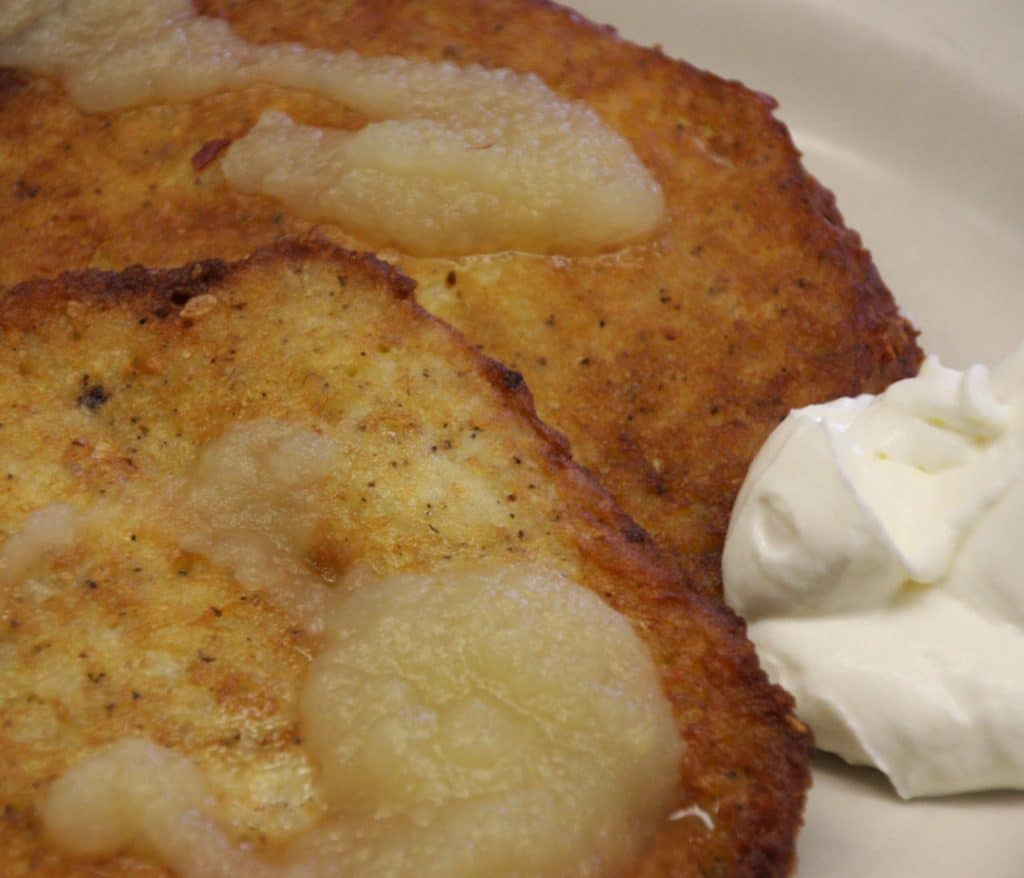 A close-up view of golden-brown German potato pancakes (Reibekuchen/Kartoffelpuffer) served on a white plate. The fried pancakes are topped with dollops of applesauce and served with a side of white sour cream.