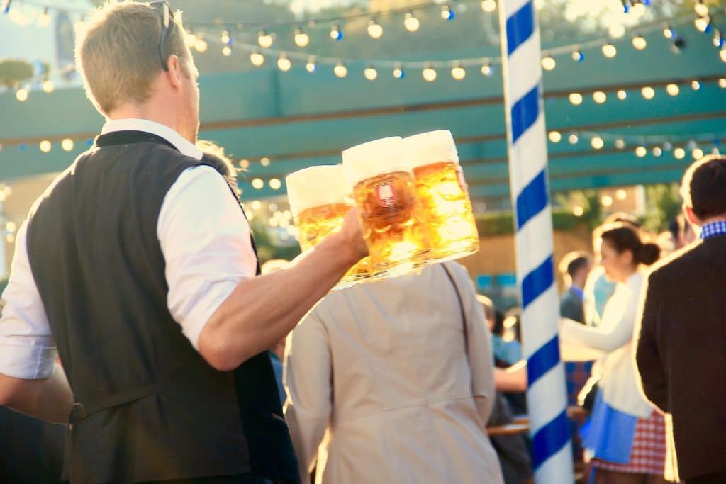 A waiter in a traditional black vest carries several one-liter Spaten beer mugs (Masskrug) filled with golden lager at the Oktoberfest festival. The scene is backlit by warm sunlight, highlighting the beer's amber color, with a festive crowd and a blue-and-white Bavarian pole in the blurred background.