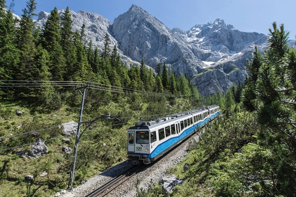 A blue and white Bayerische Zugspitzbahn cogwheel train ascending a track through a pine forest, with towering rocky Alpine peaks in the background under a clear blue sky.