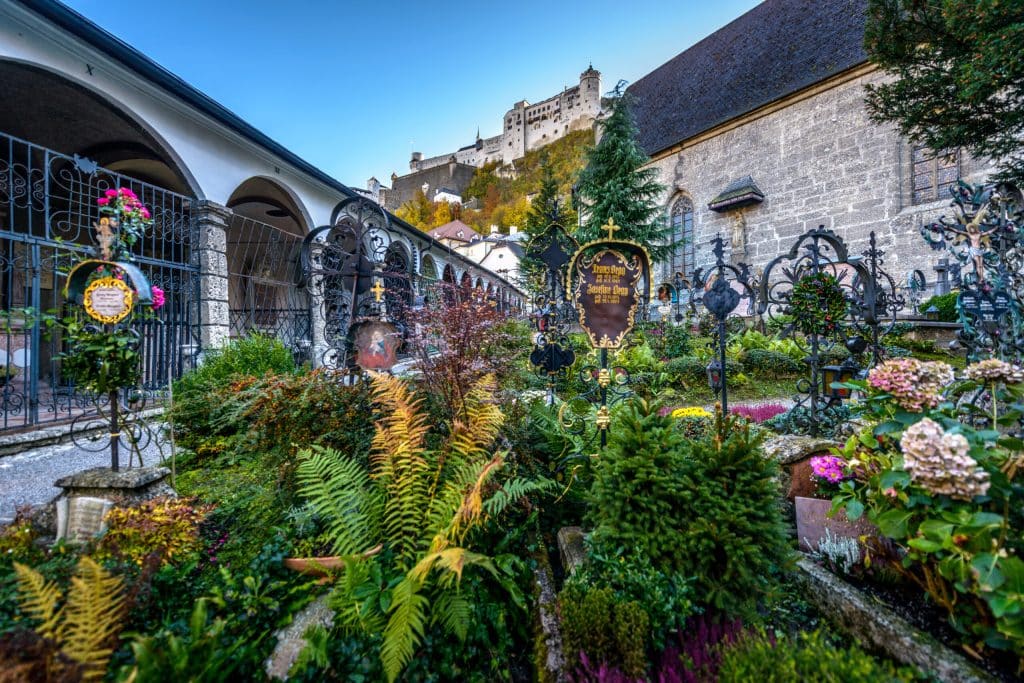 A low-angle view of St. Peter's Cemetery (Petersfriedhof) in Salzburg, filled with ornate wrought-iron grave crosses and lush flowers. The Hohensalzburg Fortress stands majestically on the hill in the background against a blue sky.