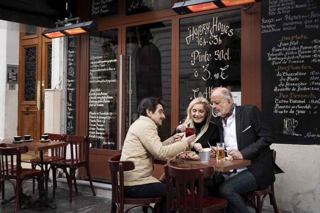 Three older adults sitting at a wooden table on an outdoor French bistro terrace, smiling while looking at a smartphone screen. The background features a window with "Happy Hours" text and a chalkboard menu listing dishes like "Faux Filet" and "Nos Planches."