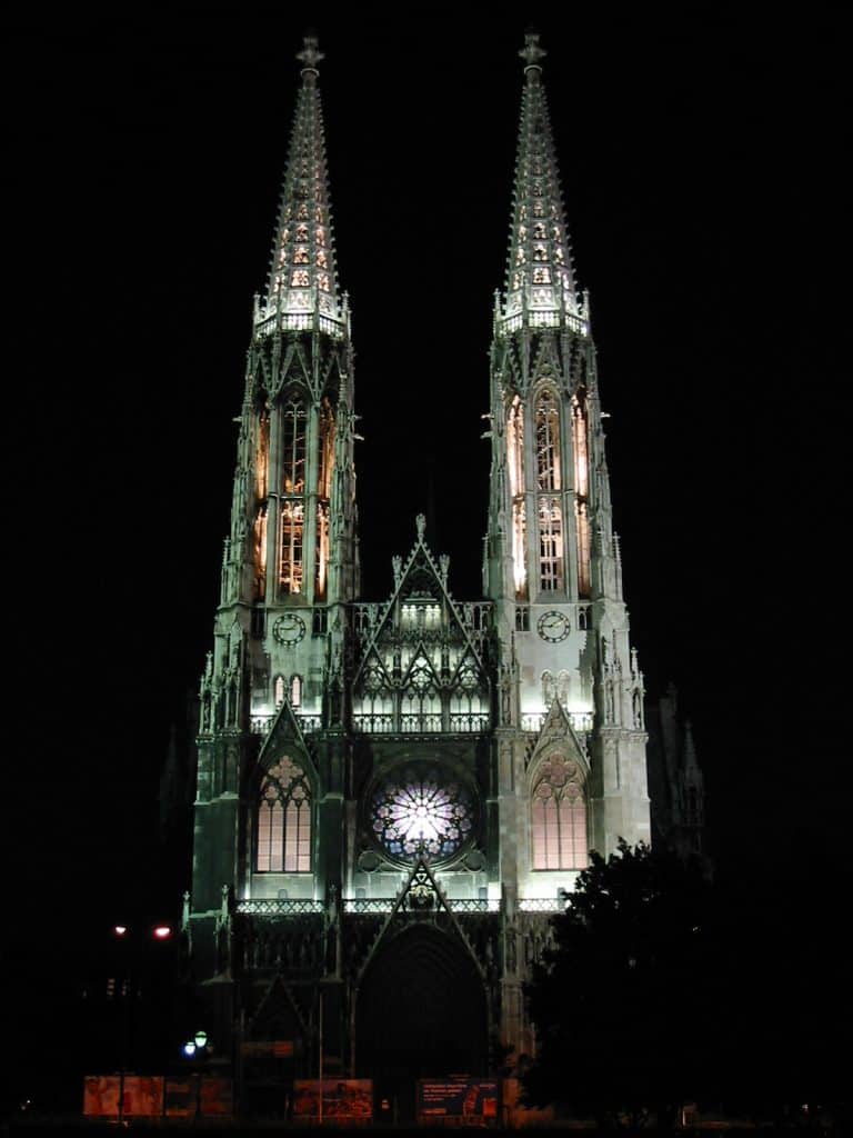 Low-angle night photography of the Votivkirche in Vienna, featuring illuminated twin neo-Gothic spires and a central rose window against a solid black sky.