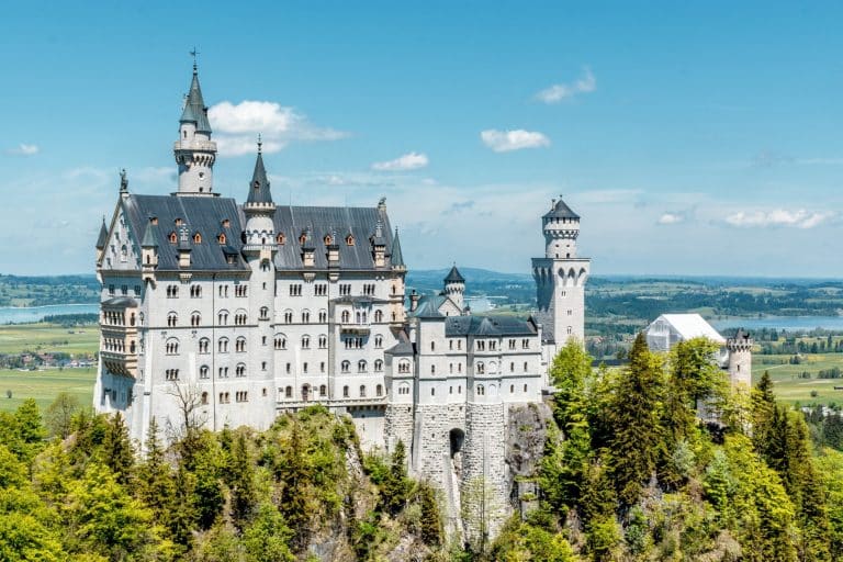 A majestic landscape view of Neuschwanstein Castle perched on a rugged, forest-covered hill in Bavaria, Germany. The white limestone castle with its iconic dark blue turrets stands against a backdrop of distant lakes and a bright blue sky with scattered clouds.