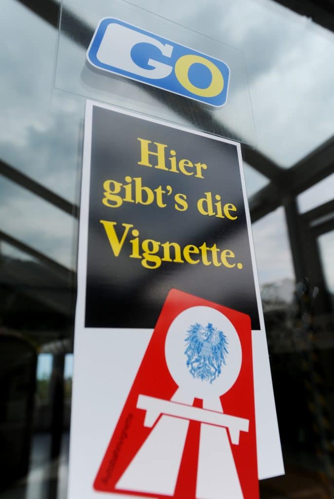 A close-up of a sign on a glass door reading 'Hier gibt's die Vignette' (Vignette available here). It features the yellow and blue 'GO' toll logo and a red icon showing the Austrian eagle coat of arms on a motorway symbol.