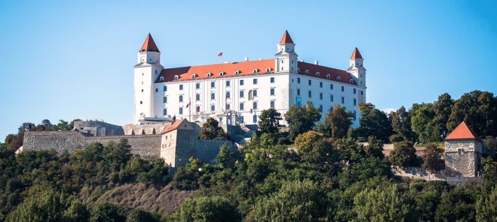 A panoramic view of the historic white Bratislava Castle (Bratislavský hrad) in Slovakia, featuring four corner towers with red roofs, situated atop a rocky hill surrounded by green trees and stone fortification walls under a clear blue sky.