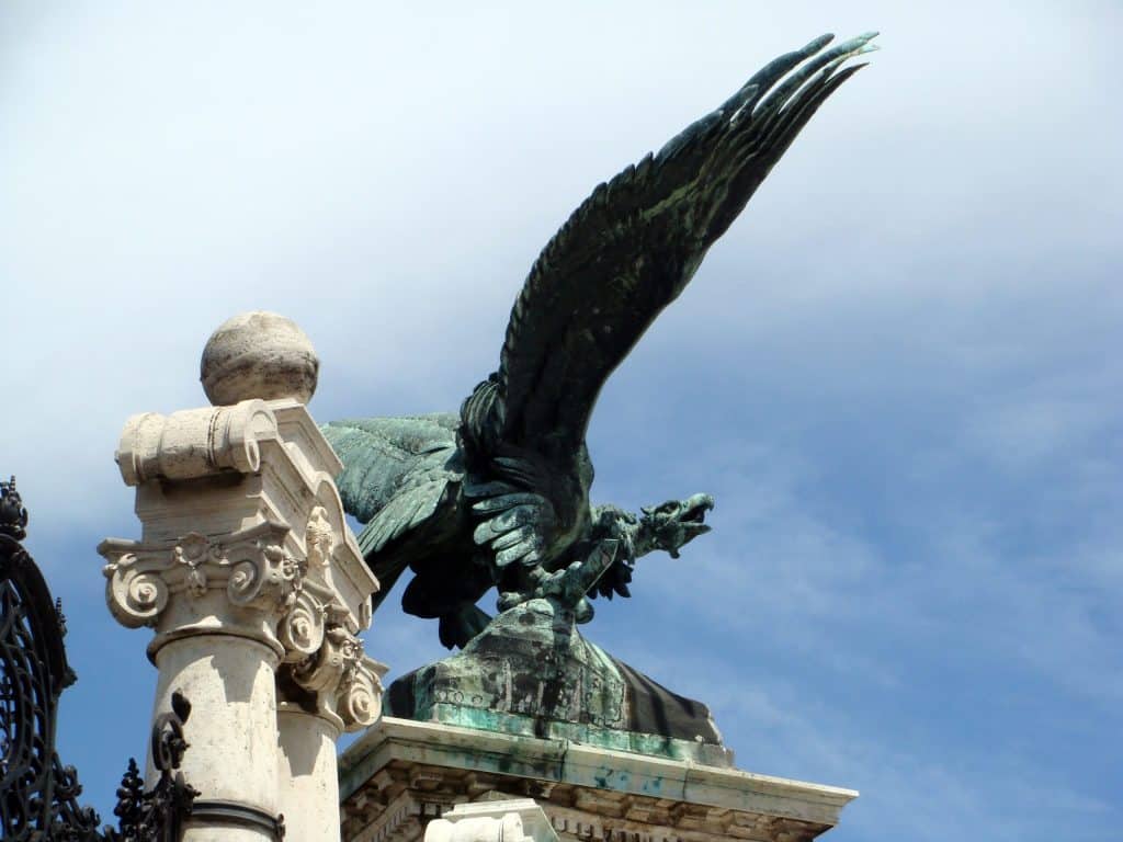 Buda Castle District: A Journey Through Hungarian History - Low-angle view of the majestic bronze Turul bird statue with wide-spread wings, perched on a stone pedestal next to an ornate limestone column at the Habsburg Gate in Buda Castle, Budapest.