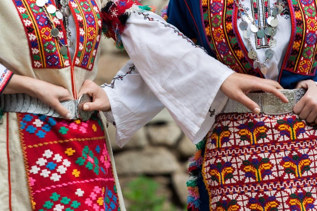 A close-up shot of two people wearing traditional Bulgarian folk costumes, featuring intricate colorful embroidery in red, blue, and yellow patterns. They are wearing silver belt buckles (pafti) and silver coin necklaces. Their hands rest on their belts.