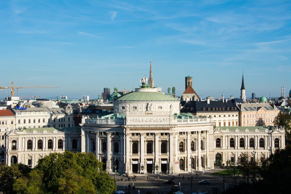 Wide shot of the Burgtheater in Vienna on a sunny day, showing the white classical facade, green copper dome, and "K.K. HOFBURGTHEATER" inscription.