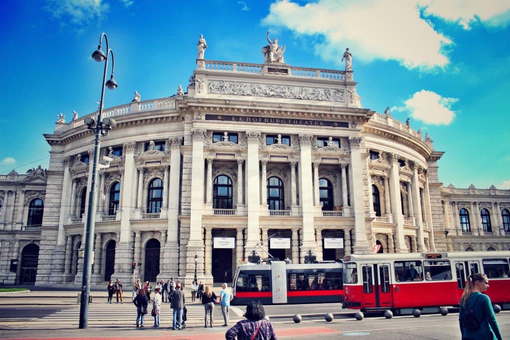 The ornate facade of the K.K. Hofburgtheater in Vienna under a blue sky, with a red and white electric tram passing by in the foreground and pedestrians waiting at a crosswalk.