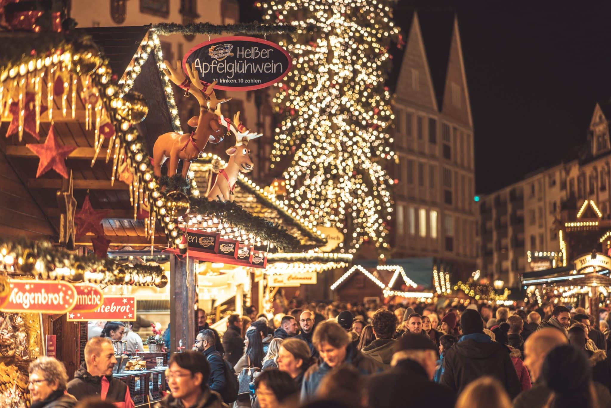 A bustling scene at a German Christmas Market (Weihnachtsmarkt) at night, featuring a wooden stall selling "Heißer Apfelglühwein" decorated with illuminated reindeer, with a large crowd and a glowing Christmas tree in the background.