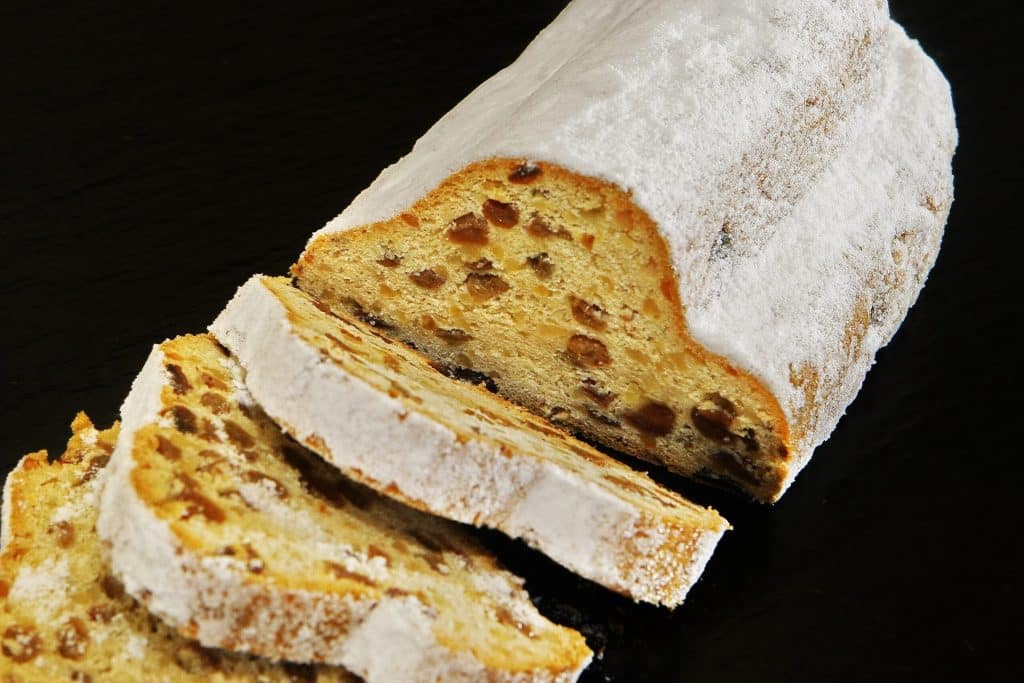 A loaf of traditional German Christmas Stollen (Christstollen) sitting on a dark surface. The bread is thickly coated in white powdered sugar. Several slices have been cut, revealing a dense golden crumb filled with raisins and candied fruit pieces.