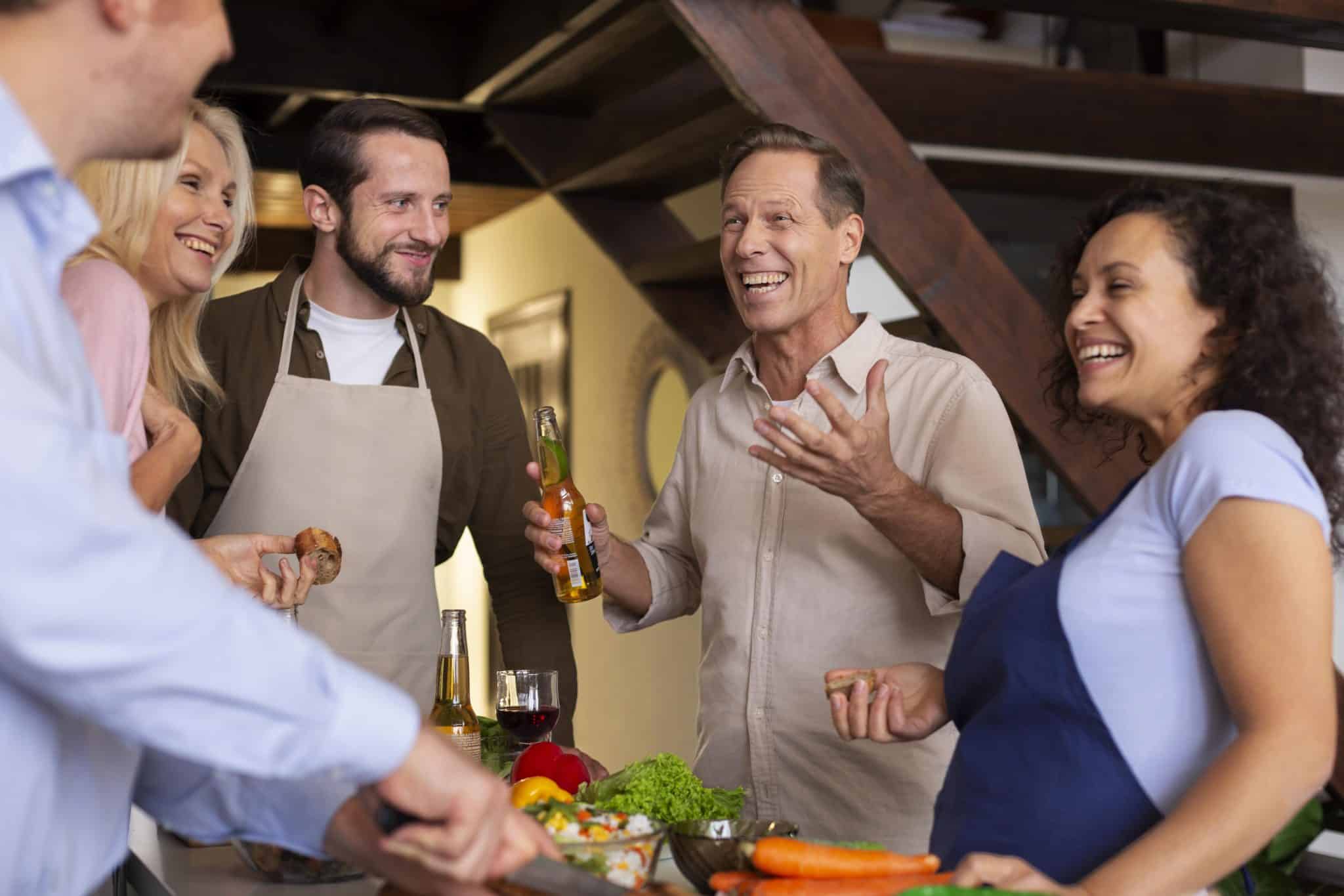 A diverse group of five happy adults laughing and talking in a modern kitchen while preparing a meal. Two men are wearing beige aprons, and a woman wears a blue apron. In the foreground, hands are chopping carrots and vegetables, while a man in the center holds a beer bottle and gestures during a conversation.