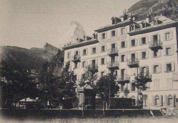 A vintage sepia-toned photograph of the Hotel Monte Rosa in Zermatt, showing the multi-story facade with balconies and shutters, with the peak of the Matterhorn faintly visible in the background.