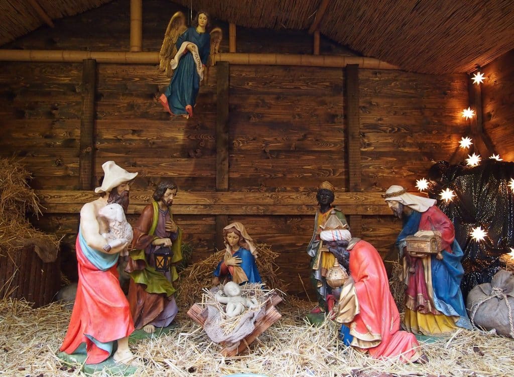 A traditional wooden Nativity scene (Krippe) inside a stable with a straw floor. Painted figures of Mary, Joseph, and the Three Wise Men surround Baby Jesus in the manger. A shepherd with a sheep stands to the left, and an angel holding a "Gloria" scroll hangs from the thatched roof.