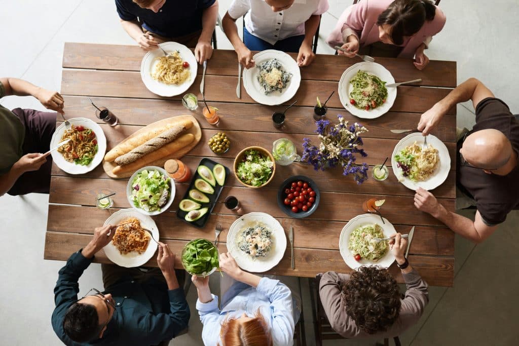 Solo Travel on Coach Tours: Single Supplements and Roommate Matching - Top-down view of eight diverse people sitting around a large rustic wooden table enjoying a pasta meal together. The table is set with various pasta dishes, salad, avocados, baguettes, drinks, and a centerpiece of dried flowers.