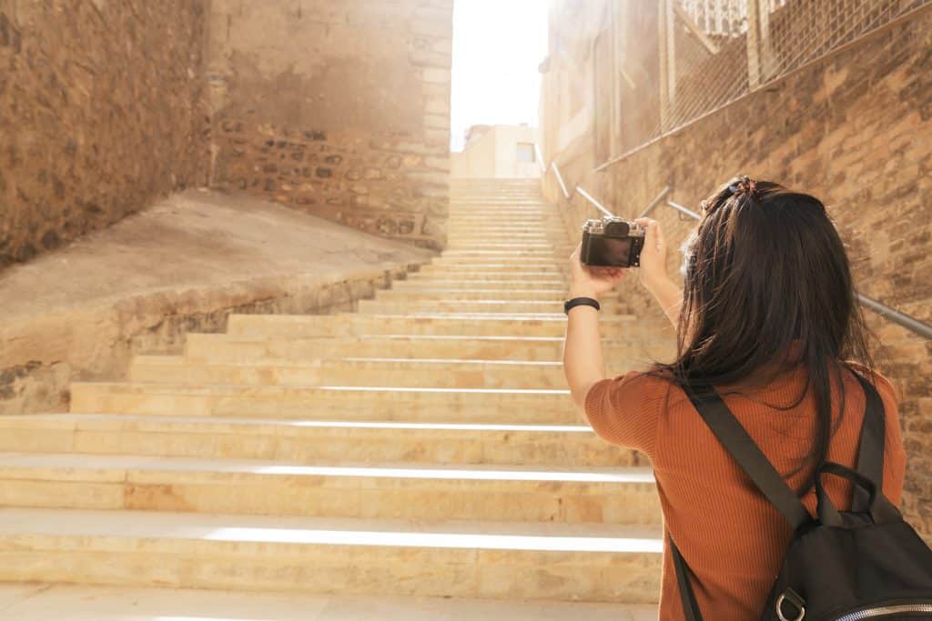 Rear view of a female tourist with a black backpack holding a camera, standing at the bottom of sunny stone steps in a historic alleyway in Cartagena, Spain.