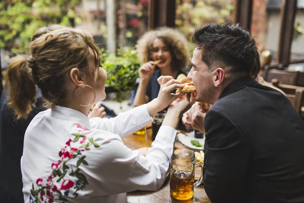 A woman with a blonde ponytail and floral embroidered shirt feeds a bite of a burger to a smiling man in a black blazer at a wooden restaurant table. In the blurred background, another woman with curly hair eats french fries. Large beer mugs sit on the table.