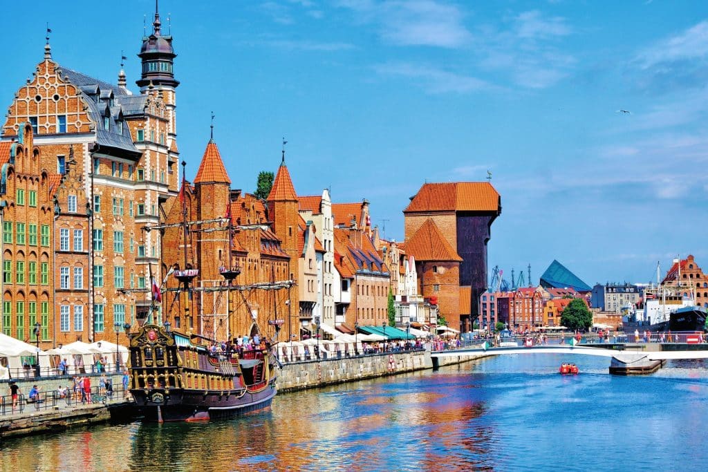 A sunny panoramic view of the Long Embankment along the Motława River in Gdańsk, Poland. The image features historic red-brick Dutch Mannerist architecture, the iconic medieval Crane (Żuraw), and a replica pirate galleon ship docked near a white pedestrian footbridge.