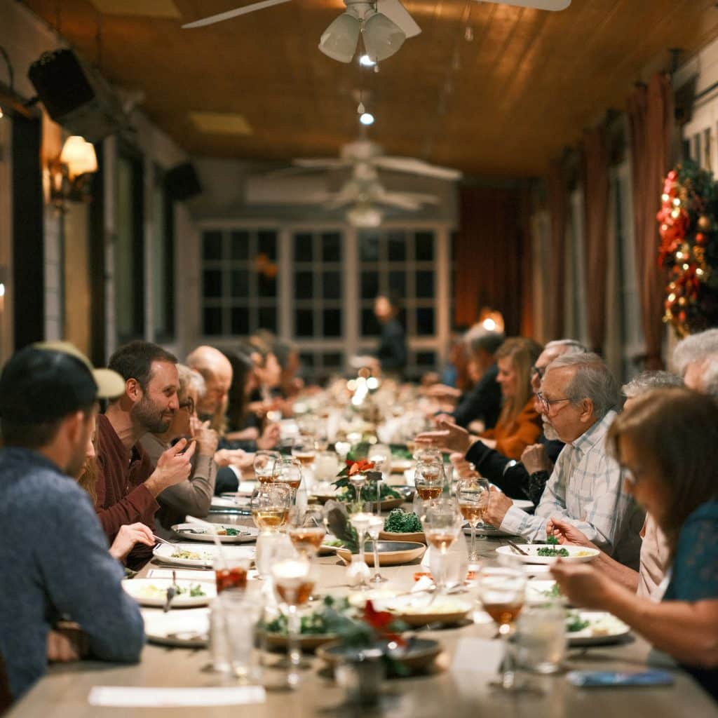A warm and candid shot of a large group of friends and family seated at a long wooden dining table on a covered porch. Guests are engaged in conversation, eating, and drinking wine under ceiling fans. A Christmas wreath is visible in the background, suggesting a holiday gathering.