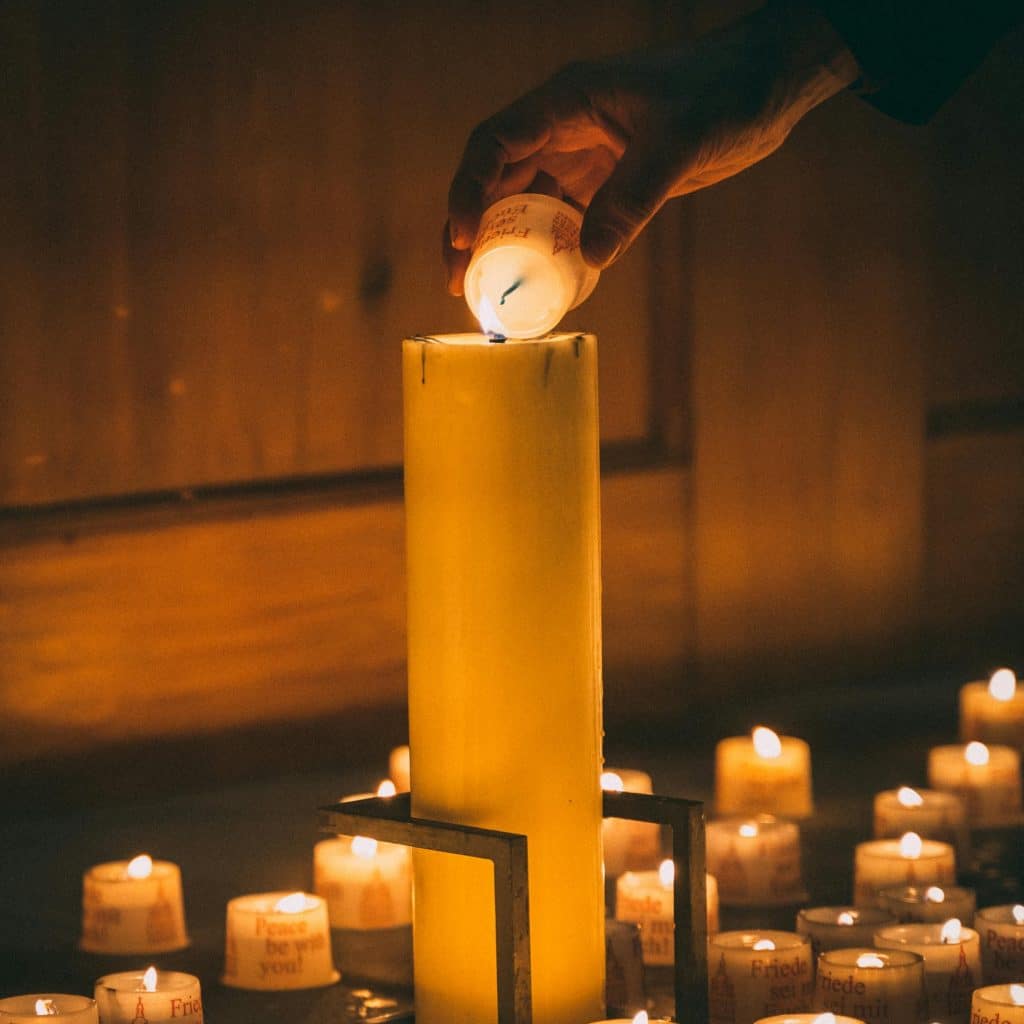 Religious Customs and Church Etiquette: A Traveler's Guide to Europe's Sacred Spaces - A close-up of a hand tilting a small glass votive candle to light the wick of a tall, thick yellow pillar candle in a dark setting. The large candle is surrounded by dozens of smaller tea lights on the floor, some printed with the text "Peace be with you!" and "Friede sei mit euch". The scene is illuminated by a warm, orange candlelight glow.