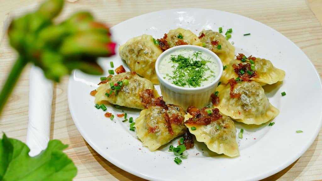 A white plate featuring a circle of homemade pierogi or varenyky dumplings topped with caramelized onions and chopped chives. A small ramekin of white dipping sauce garnished with dill sits in the center.