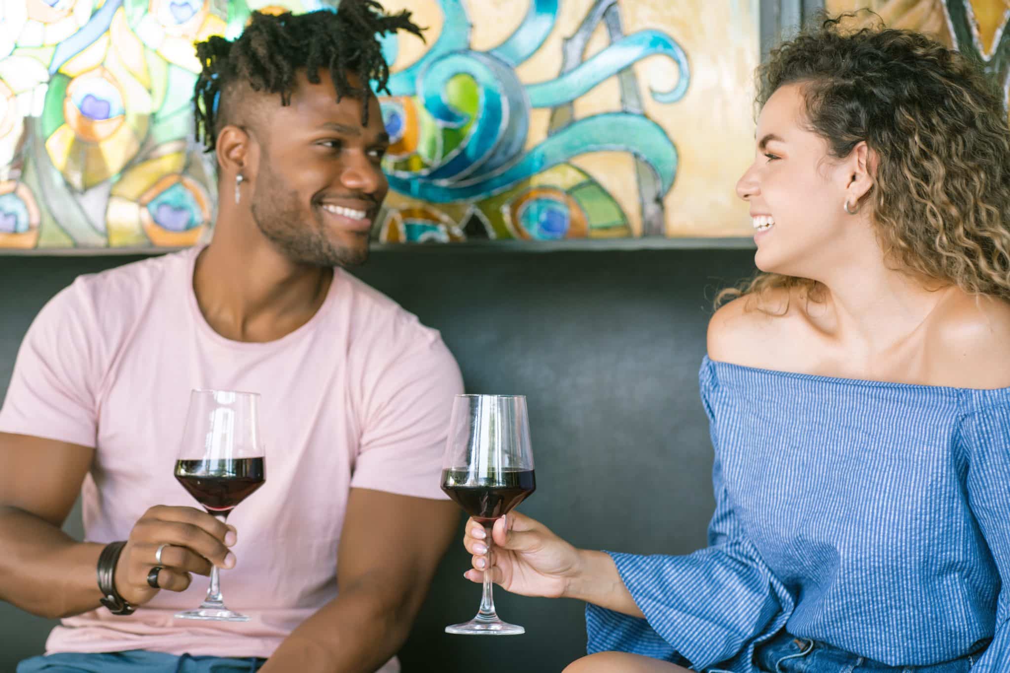 A happy young interracial couple sitting together in a restaurant with a colorful stained-glass background, holding glasses of red wine and smiling affectionately at each other during a romantic date.