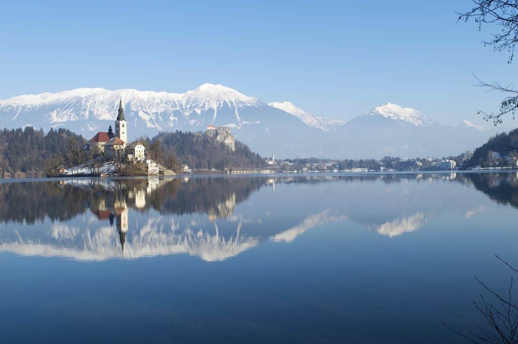 A wide panoramic view of Lake Bled in Slovenia during winter. The image features the Pilgrimage Church of the Assumption on the small island to the left and Bled Castle perched on a cliff in the distance. The snow-capped Julian Alps are perfectly reflected in the calm, mirror-like blue water under a clear sky.