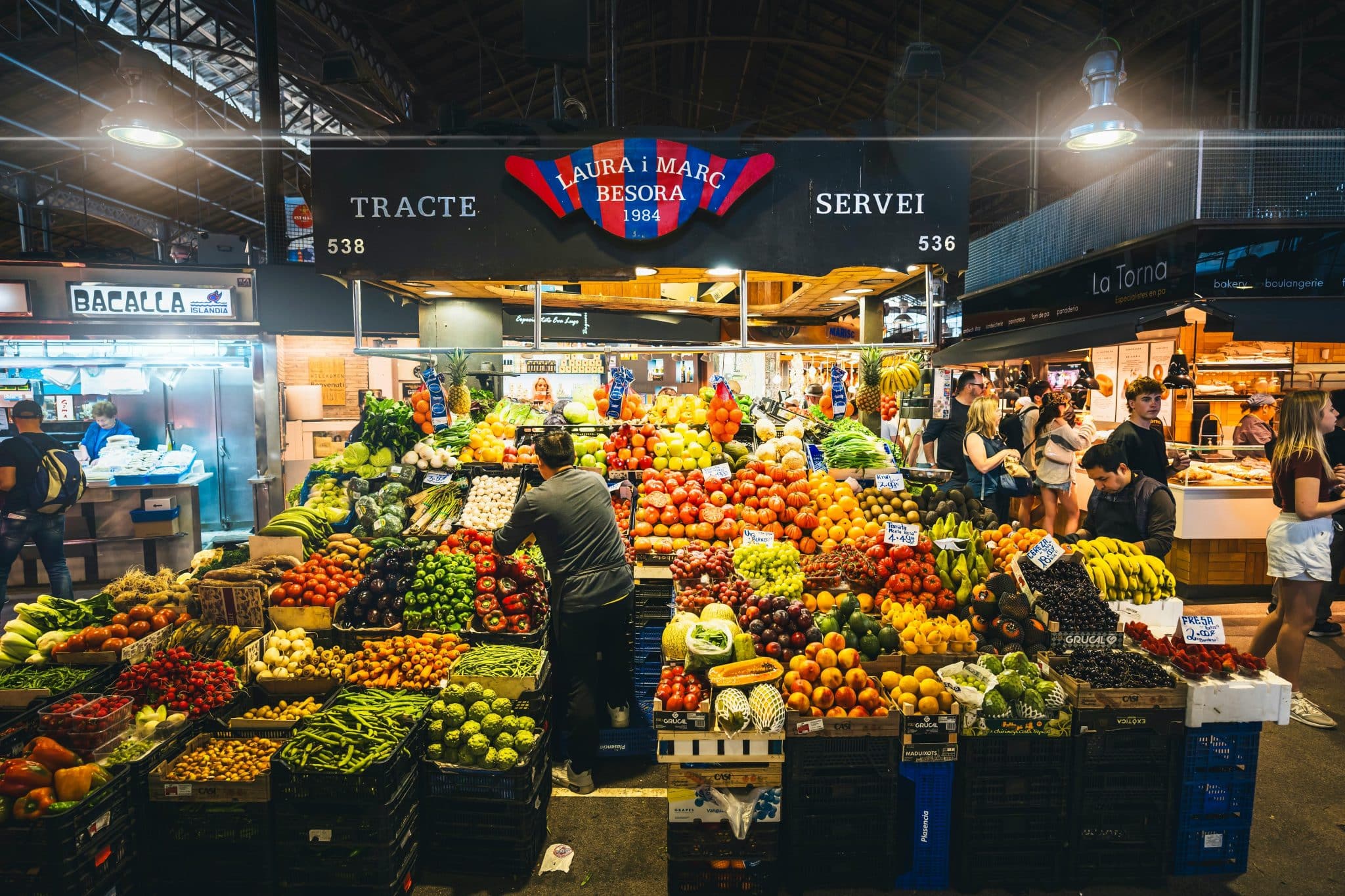 Discover the art of European market shopping, A vibrant fruit and vegetable stall named "Laura i Marc Besora" at the famous La Boqueria market in Barcelona. The stand is overflowing with fresh produce including tomatoes, peppers, artichokes, and bananas. A worker in a grey shirt arranges the display while customers browse in the background under the market's iron roof structure.