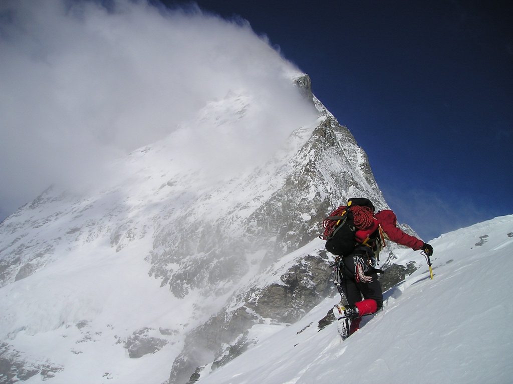 A mountaineer in a red jacket ascending a steep snowy ridge on the Matterhorn, equipped with an ice axe, crampons, and a coiled red rope, with spindrift blowing off the peak against a deep blue sky.