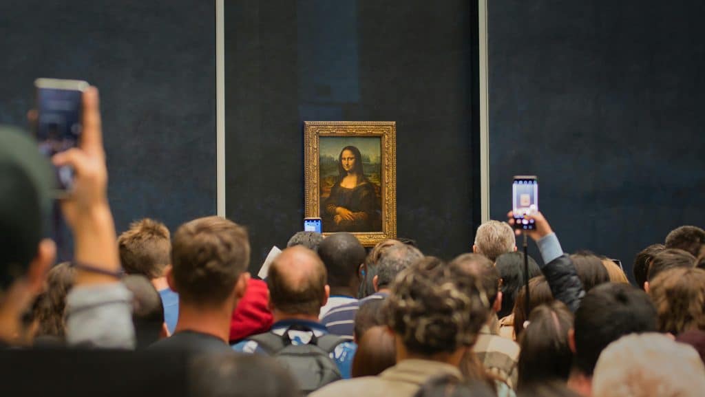 How to Decode a European Coach Tour Brochure: A Tour Director's Guide to Finding the Right Fit - A large crowd of tourists viewed from behind standing in front of the Mona Lisa painting at the Louvre Museum, with many people holding up smartphones to take photos of the artwork on the dark blue wall.
