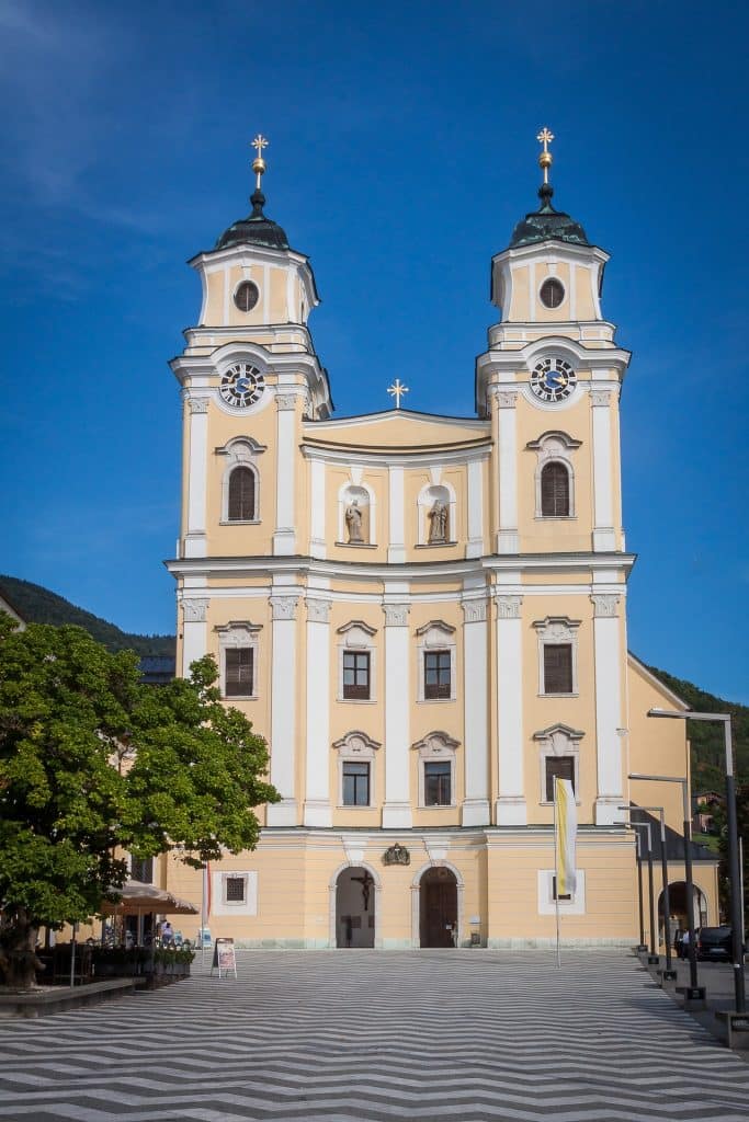 The grand yellow and white Baroque facade of the Basilica of St. Michael in Mondsee, Austria, featuring twin towers with dark domes and a public square with a zigzag pavement pattern in the foreground.