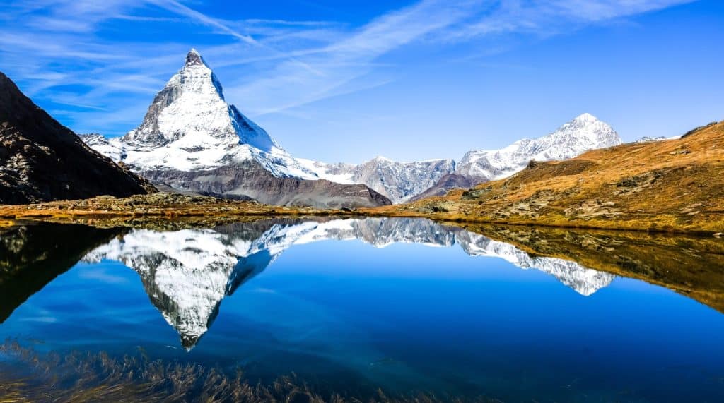 A vibrant landscape photo of the Matterhorn reflected perfectly in the calm blue waters of Riffelsee lake on a sunny day.