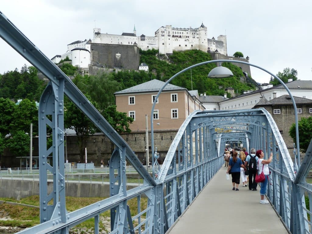 Tourists walking across the blue steel Art Nouveau Mozartsteg bridge in Salzburg, Austria, with the historic white Hohensalzburg Fortress sitting atop a green hill in the background. A pedestrian is seen wearing a backpack with a FILA logo.