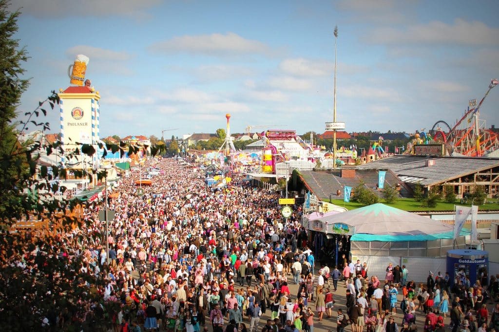 A massive, dense crowd fills the main avenue of the Oktoberfest festival grounds in Munich on a sunny day. The iconic Paulaner tower with a giant beer mug sits on the left, flanked by carnival rides, beer tents, and a blue ATM container labeled "Geldautomat" in the foreground.