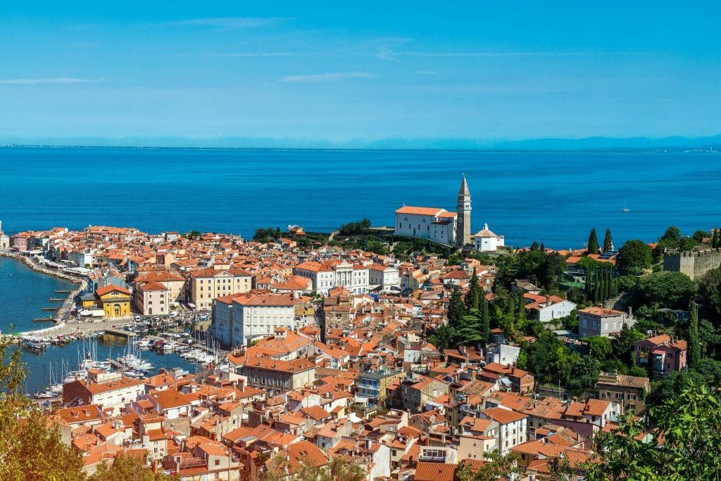 A high-angle, panoramic view of the coastal town of Piran, Slovenia, featuring a dense cluster of historic buildings with terracotta orange roofs, the prominent bell tower of St. George's Parish Church on the ridge, a marina filled with small white boats, and the calm blue Adriatic Sea extending to the distant mountains on the horizon.