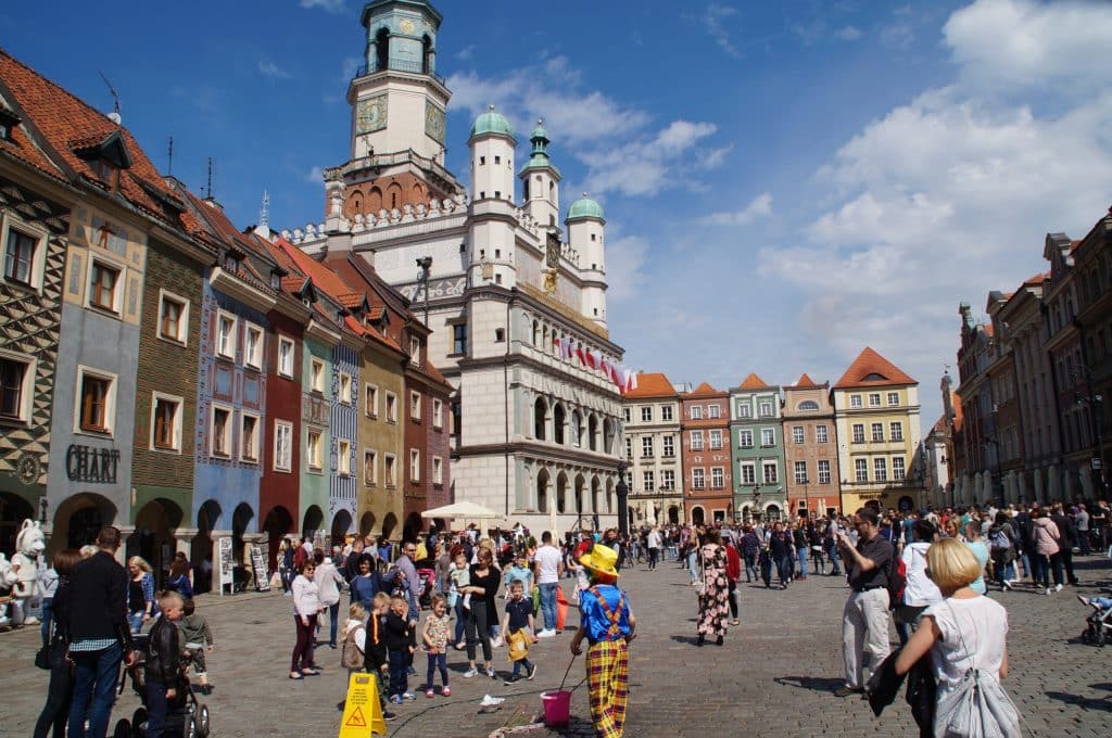 Poznań Old Market Square: A Guide to the Goats, Ghosts, and Croissants of Poland's Birthplace - 
A sunny daytime view of the Old Market Square (Stary Rynek) in Poznań, Poland, featuring the Renaissance Town Hall with its clock tower and colorful merchant houses. A crowd of tourists and locals fills the cobblestone square, including a street performer dressed as a clown in the foreground.