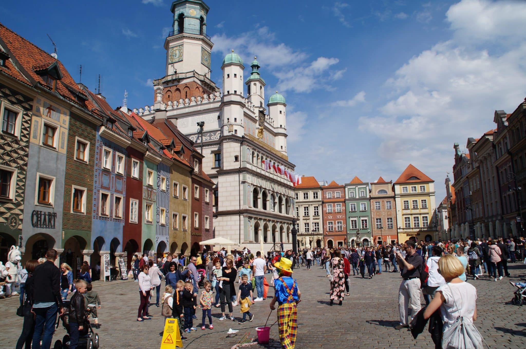 A sunny daytime view of the Old Market Square (Stary Rynek) in Poznań, Poland, featuring the Renaissance Town Hall with its clock tower and colorful merchant houses. A crowd of tourists and locals fills the cobblestone square, including a street performer dressed as a clown in the foreground.