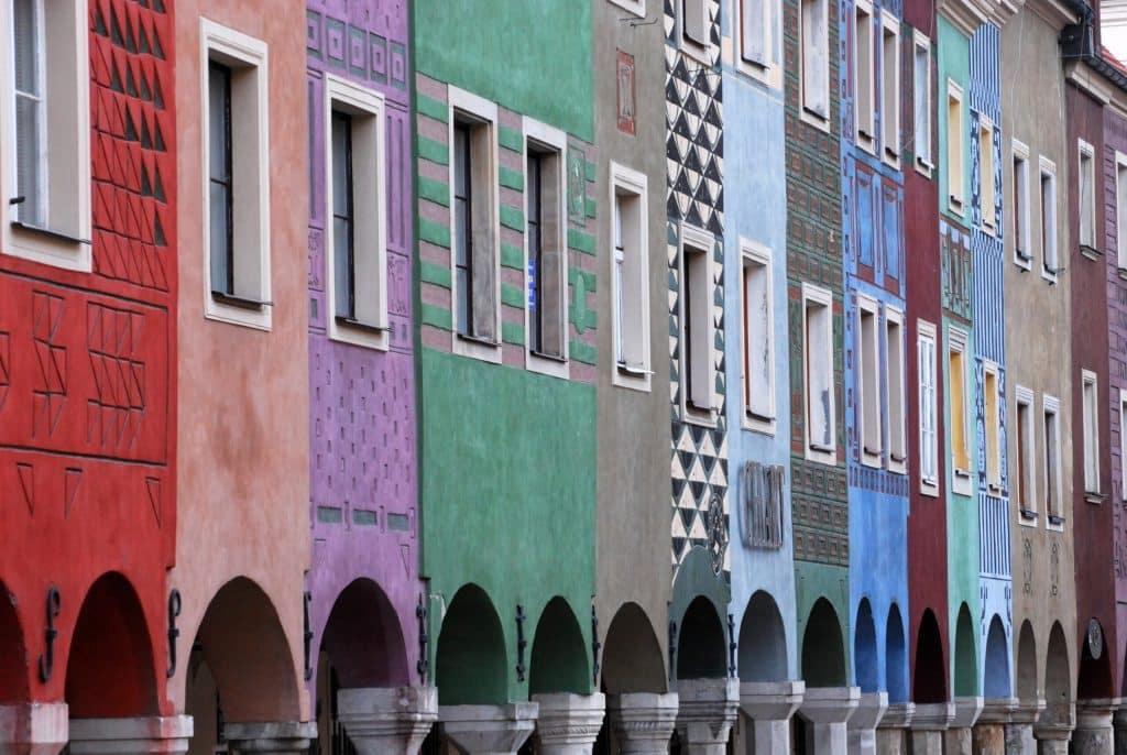 A detailed view of the colorful Merchant Houses (Domki Budnicze) in Poznań's Old Market Square. The narrow tenement buildings feature vibrant red, pink, green, and blue facades with intricate sgraffito geometric patterns and ground-floor arcades. A sign reading "CHART" is visible on the blue building.