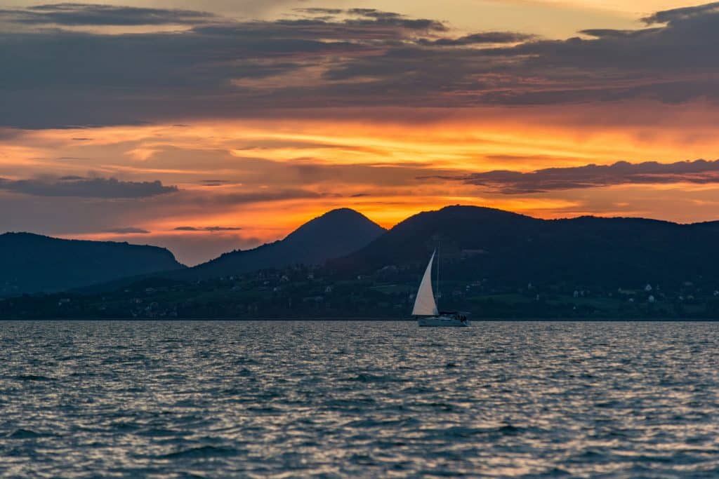 A wide shot of a white sailboat navigating a choppy lake at dusk, set against a backdrop of dark silhouetted volcanic mountains and a dramatic orange and grey cloudy sky.