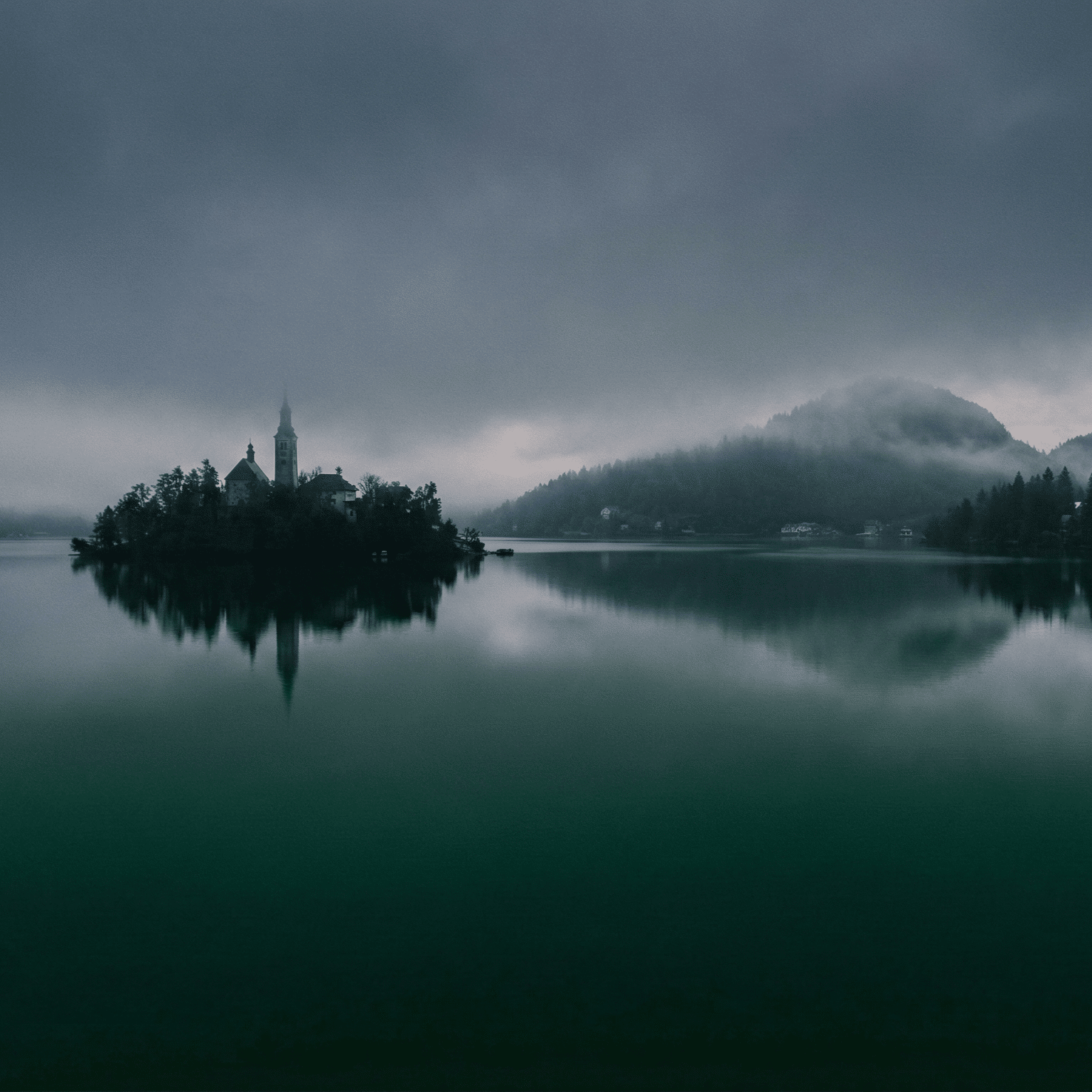 A moody, atmospheric landscape photograph of the Pilgrimage Church of the Assumption of Maria on an island in the center of Lake Bled, Slovenia. The scene is shrouded in thick fog and mist, with the dark reflection of the island visible in the calm, deep teal water, backed by silhouette mountains.