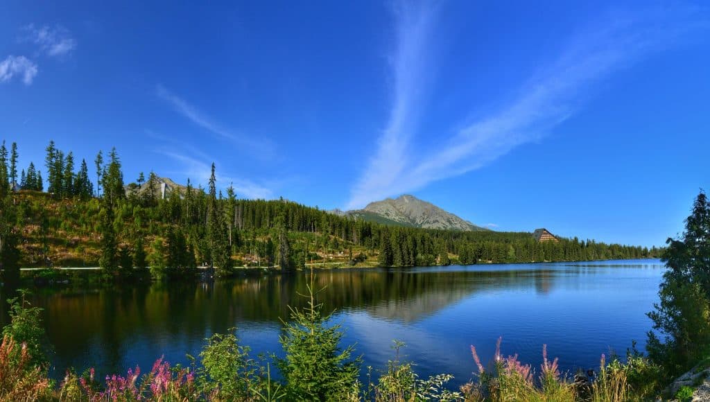 A panoramic summer view of Štrbské Pleso lake in the High Tatras, Slovakia, reflecting the deep blue sky and spruce forest, with the distinctive triangular Hotel Patria and Predné Solisko mountain peak in the background.